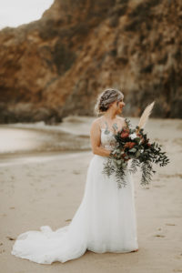 Boho bride holding wild moody bohemian bouquet on the beach in Laguna Beach, CA