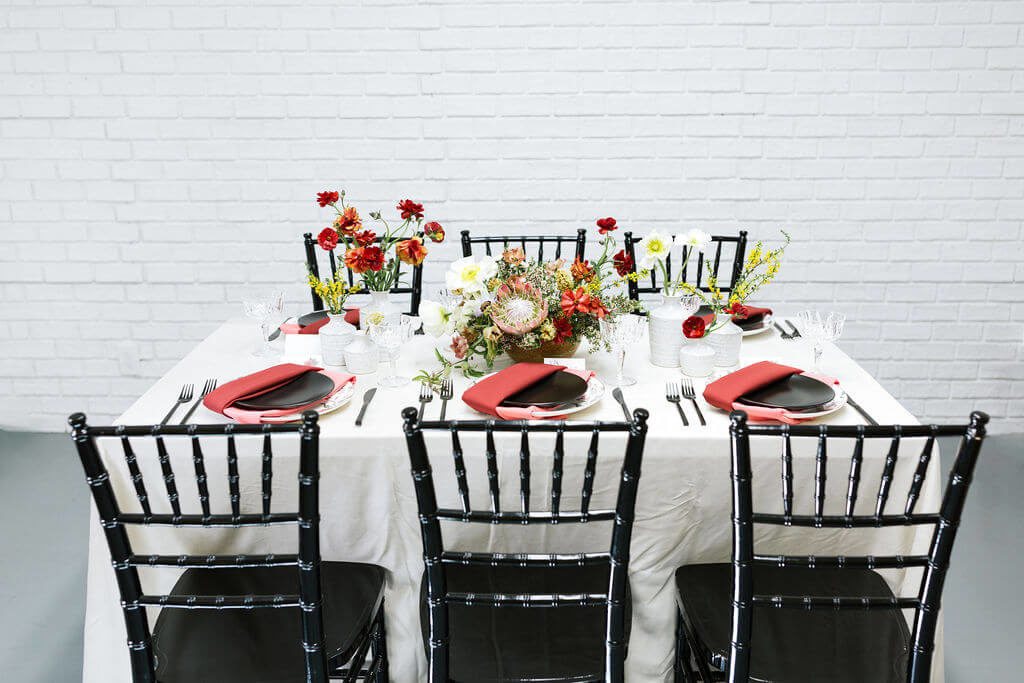 Colorful Mother's Day Brunch table with white velvet linen and black chairs. Pink and red napkins and invitations inspired by mom tattoos. Floral pattern plates are set with black matte flatware.