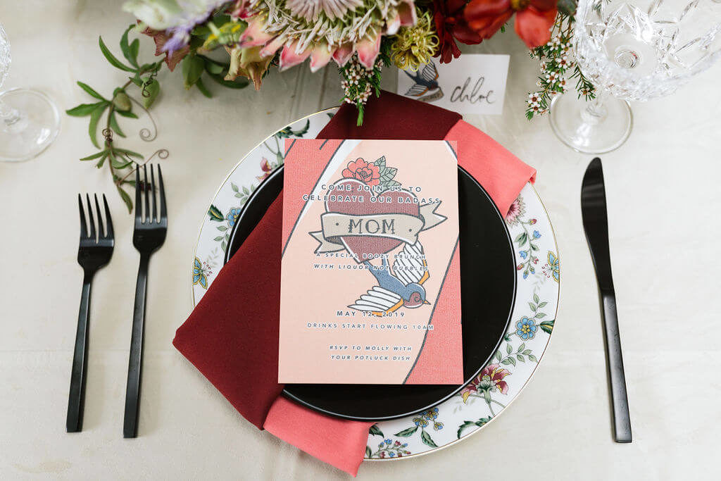 Colorful Mother's Day Brunch table with white velvet linen and black chairs. Pink and red napkins and invitations inspired by mom tattoos. Floral pattern plates are set with black matte flatware.