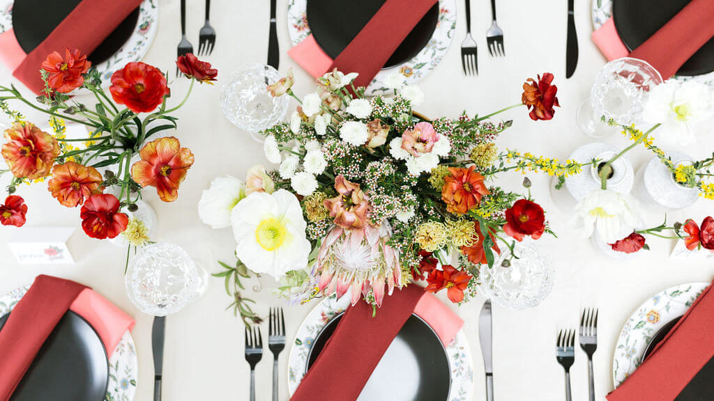 Colorful Mother's Day Brunch table with white velvet linen and black chairs. Pink and red napkins and invitations inspired by mom tattoos. Floral pattern plates are set with black matte flatware.