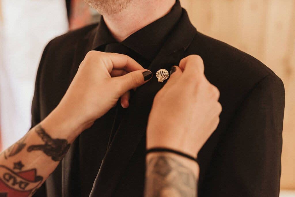 Groom getting a custom lapel pin on his suit. Groom in all black suit and shirt.