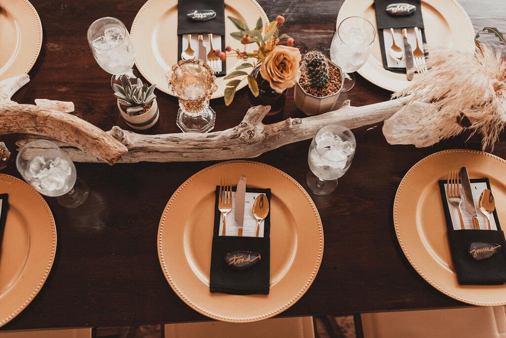 Wedding reception table with gold charger, black napkin, amber bud vases and dark flowers. Driftwood pieces on long wooden tables, pampass grass, king protea and cacti.