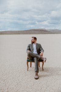 A groom poses in a chair in the middle of the desert before his boho style wedding ceremony.