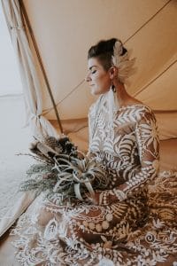 A bride with a creative feather hair piece and air plant bouquet poses in a clamping tent before her desert wedding ceremony.