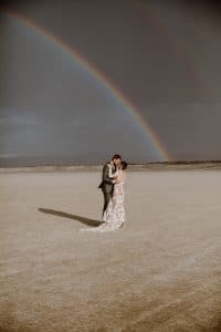 A bride and groom stand under a double rainbow in the desert for a festival style wedding inspired by Burning Man.